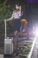 A woman standing next to a silver suitcase on a sidewalk.